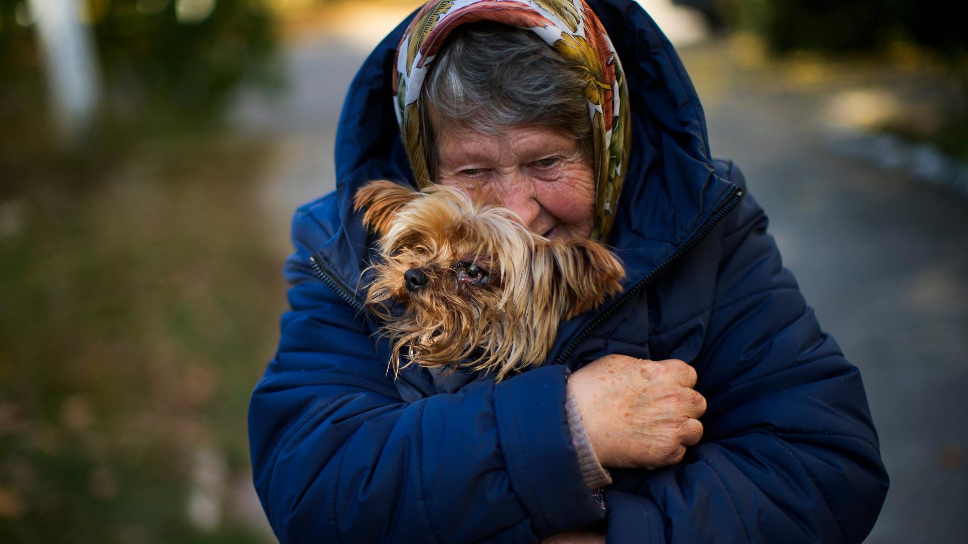 Die alte Frau in blauer Winterjacke und Kopftuch steht auf einer Straße und wärmt ihren kleinen Hund unter dem Mantel. Die alte Frau in blauer Winterjacke und Kopftuch steht auf einer Straße und wärmt ihren kleinen Hund unter dem Mantel.