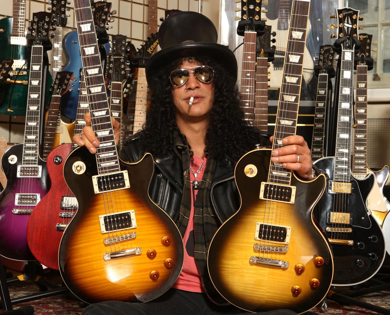 Der amerikanische Musiker Slash posiert mit zwei Gibson Gitarren. Er trägt einen schwarzen Zylinder, eine dunkle Sonnenbrille, hat schwarze lockige Haar und eine Zigarette im Mund. Der amerikanische Musiker Slash posiert mit zwei Gibson Gitarren. Er trägt einen schwarzen Zylinder, eine dunkle Sonnenbrille, hat schwarze lockige Haar und eine Zigarette im Mund.