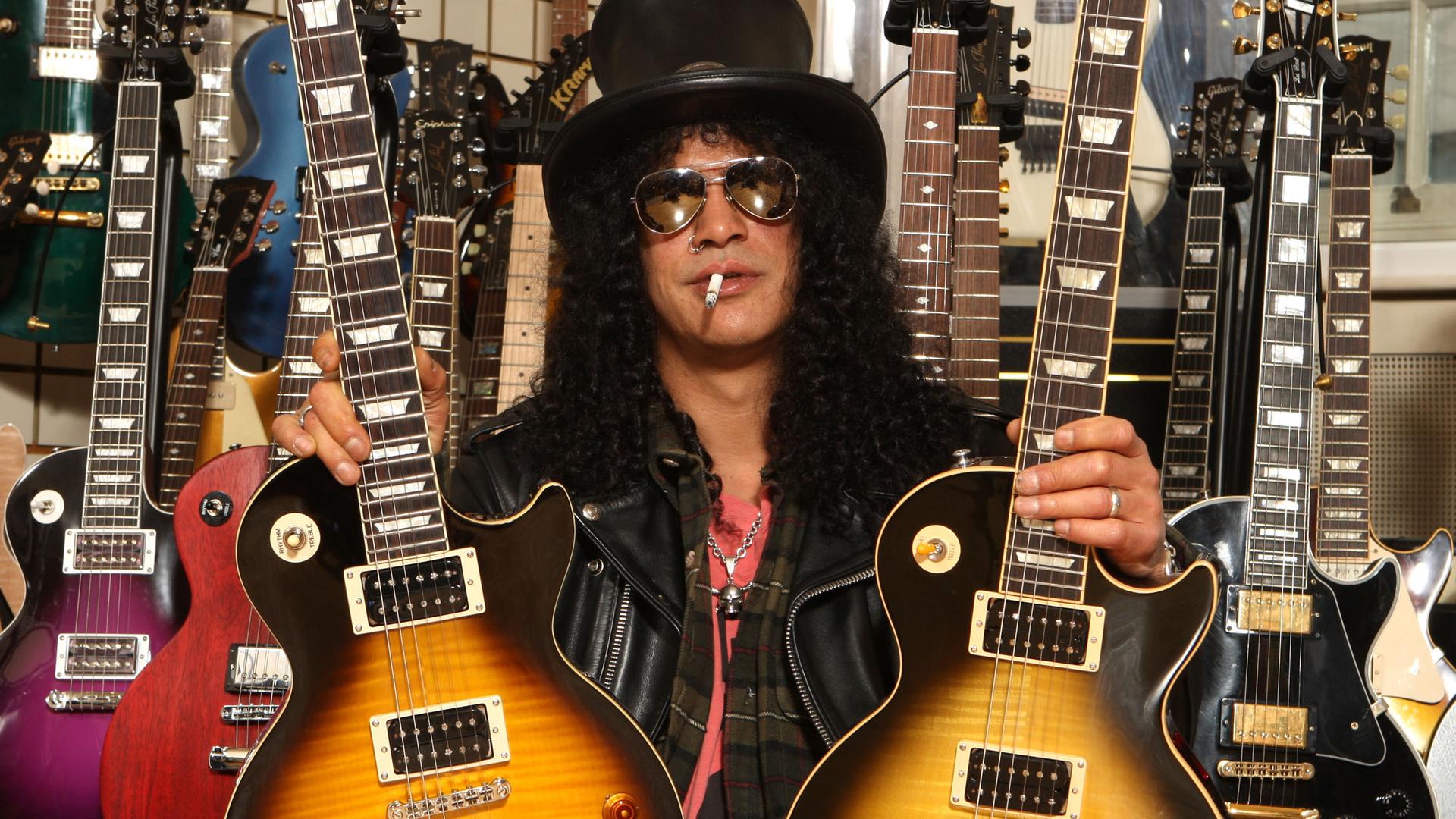 Der amerikanische Musiker Slash posiert mit zwei Gibson Gitarren. Er trägt einen schwarzen Zylinder, eine dunkle Sonnenbrille, hat schwarze lockige Haar und eine Zigarette im Mund. Foto: dpa Der amerikanische Musiker Slash posiert mit zwei Gibson Gitarren. Er trägt einen schwarzen Zylinder, eine dunkle Sonnenbrille, hat schwarze lockige Haar und eine Zigarette im Mund. Foto: dpa