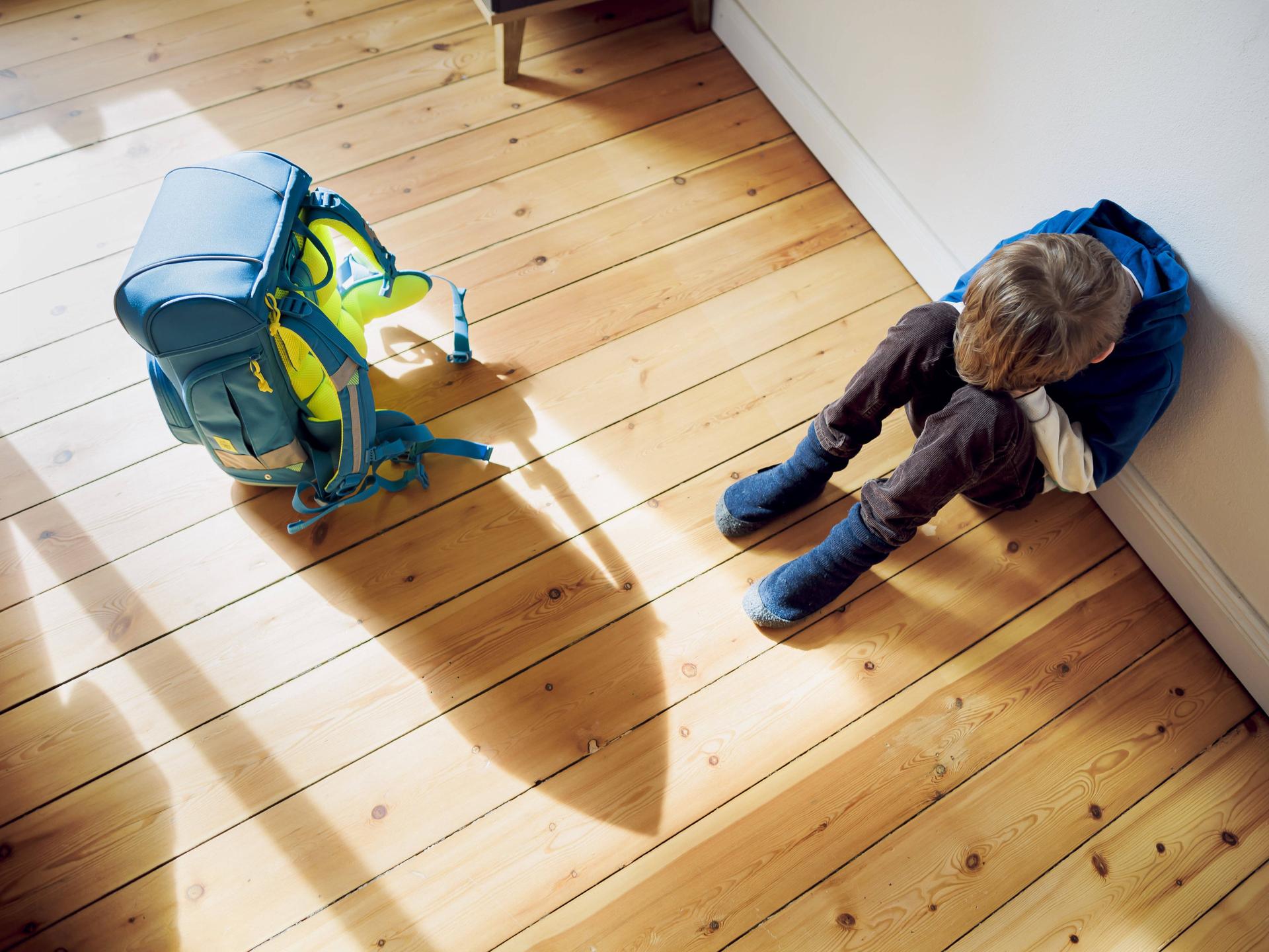 Ein Kind sitzt auf dem hölzernen Fußboden, die Beine angewinkelt, das Gesicht in den Händen vergraben. Neben ihm steht der Schulranzen. Er wirft einen langen Schatten auf den Boden.
