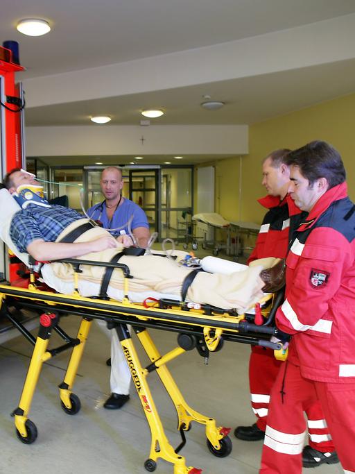 Ein Patient wird aus einem Rettungswagen auf einer Bahre von zwei Männern in roten Overalls in die Notaufnahme eines Krankenhauses gefahren.