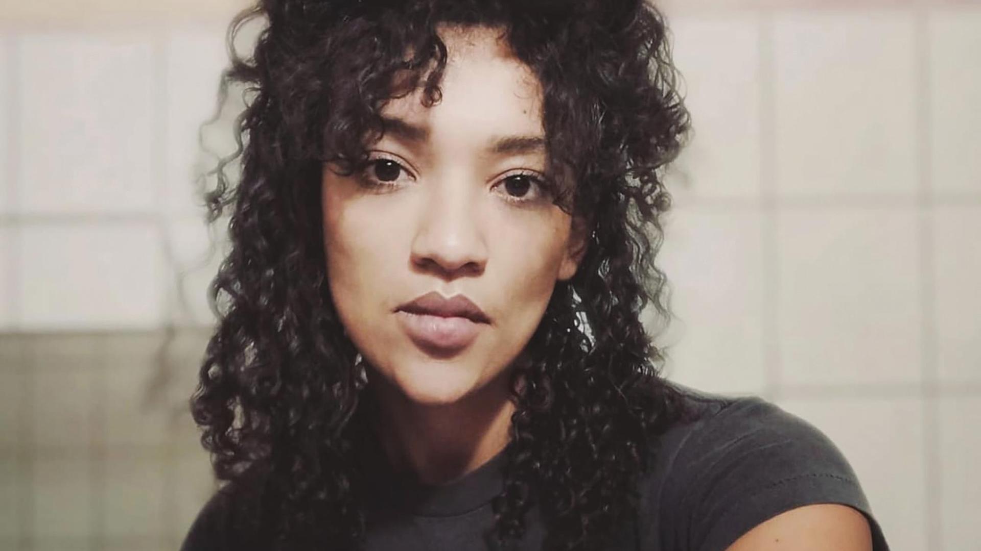 Eine Frau mit langen schwarzen Locken blickt direkt in die Kamera. Sie trägt ein schwarzes T-Shirt und sitzt vor einer gekachelten Wand. Eine Frau mit langen schwarzen Locken blickt direkt in die Kamera. Sie trägt ein schwarzes T-Shirt und sitzt vor einer gekachelten Wand.