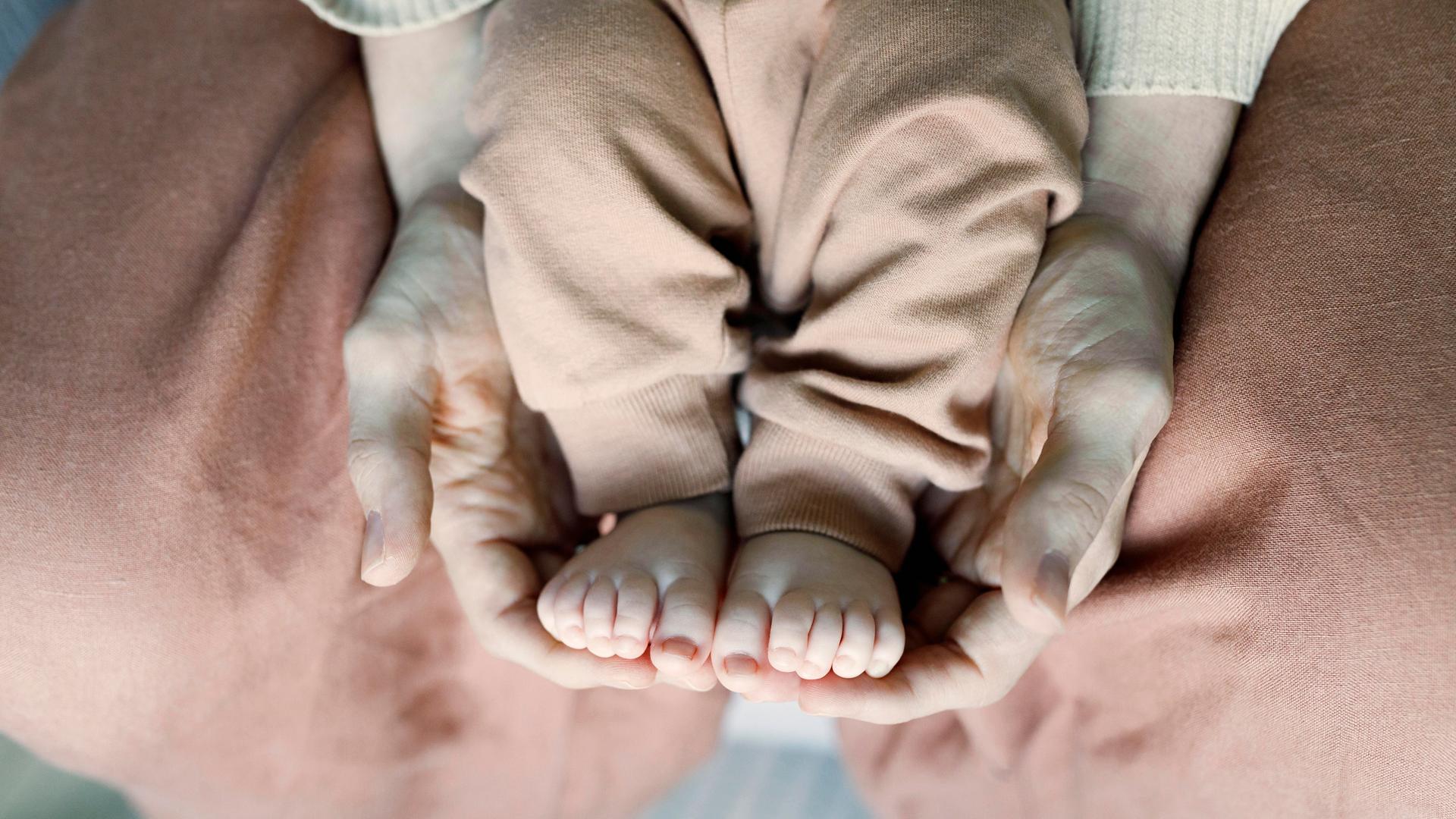 Die Füße eines Babys werden von den Händen einer erwachsenen Person auf dem Schoß gehalten. Die Füße eines Babys werden von den Händen einer erwachsenen Person auf dem Schoß gehalten.