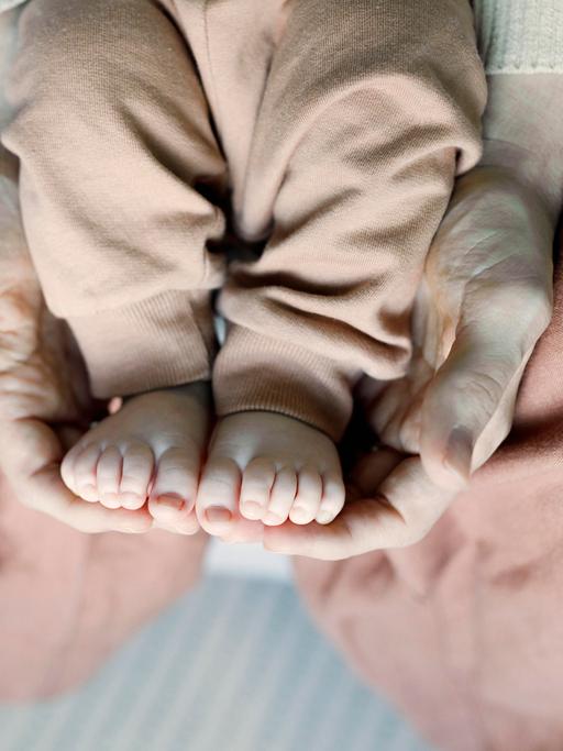 Die Füße eines Babys werden von den Händen einer erwachsenen Person auf dem Schoß gehalten.