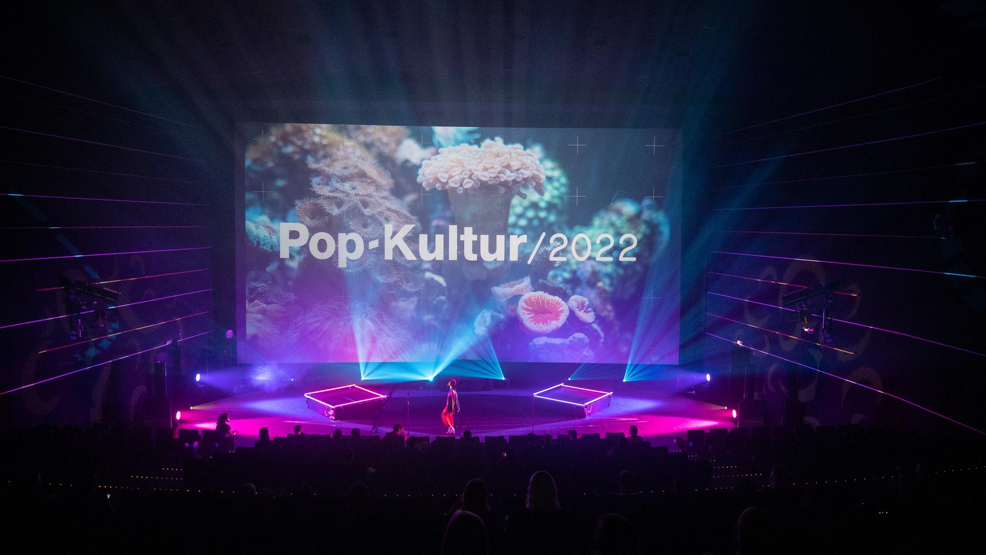 Nuha Ruby Ra steht auf der großen Bühne des Kinos in der Kulturbrauerei, die rot-lila ausgeleuchtet ist und ein Foto einer weißen Koralle im Hintergrund zeigt mit den Schriftzug "Pop-Kultur". Nuha Ruby Ra steht auf der großen Bühne des Kinos in der Kulturbrauerei, die rot-lila ausgeleuchtet ist und ein Foto einer weißen Koralle im Hintergrund zeigt mit den Schriftzug "Pop-Kultur".