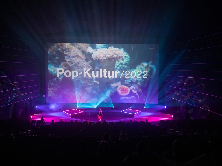Nuha Ruby Ra steht auf der großen Bühne des Kinos in der Kulturbrauerei, die rot-lila ausgeleuchtet ist und ein Foto einer weißen Koralle im Hintergrund zeigt mit den Schriftzug "Pop-Kultur".