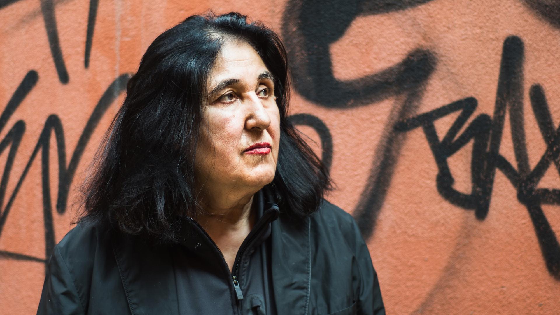 Porträt von Emine Sevgi Özdamar. Sie hat schwarze schulterlange Haare, trägt ein schwarzes Hemd und steht vor einer Wand mit Graffiti. Porträt von Emine Sevgi Özdamar. Sie hat schwarze schulterlange Haare, trägt ein schwarzes Hemd und steht vor einer Wand mit Graffiti.