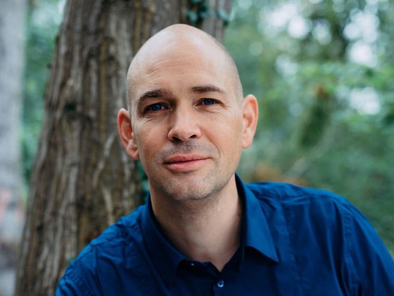 Der Berater Marc Wallert steht vor einem Baumstamm im Wald und blickt in die Kamera.