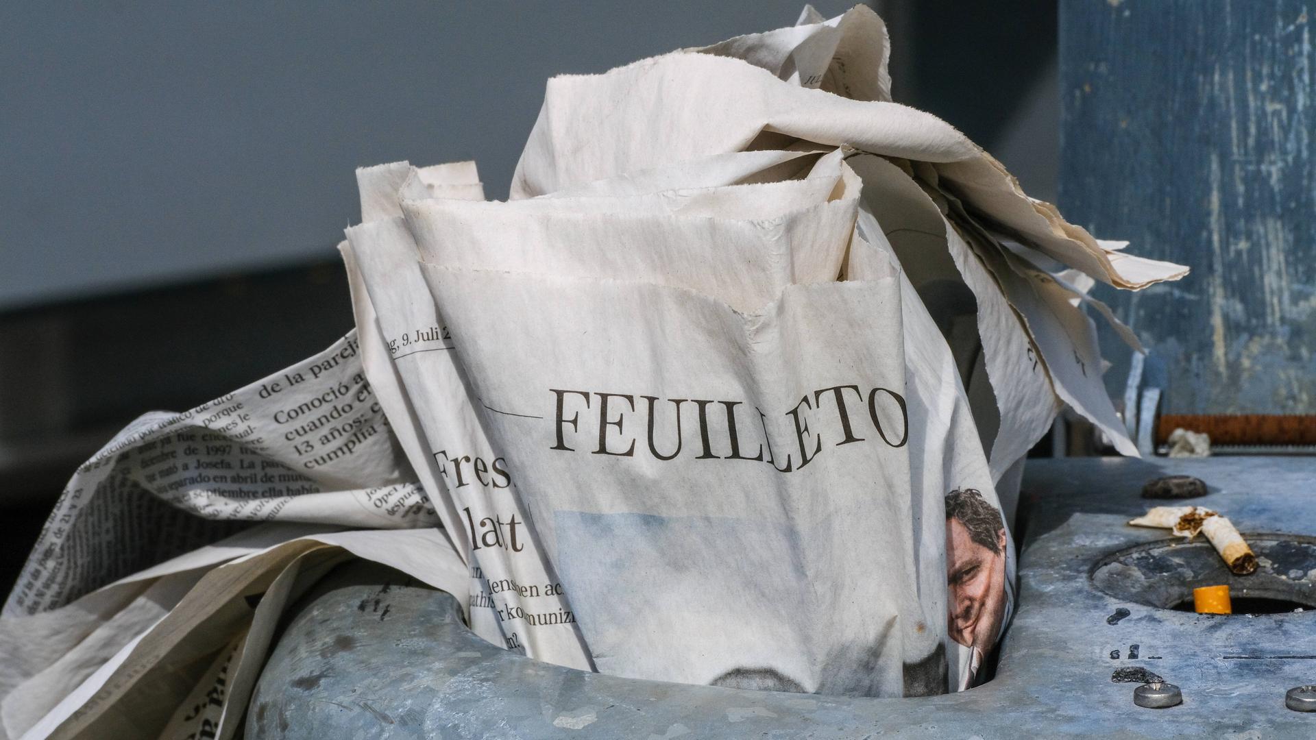 Eine gedruckte Zeitung steckt im Mülleimer einer Haltestelle. Aufgeschlagen ist das Feuilleton.