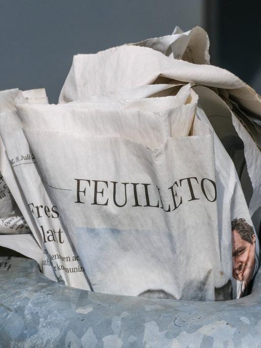 Eine gedruckte Zeitung steckt im Mülleimer einer Haltestelle. Aufgeschlagen ist das Feuilleton.