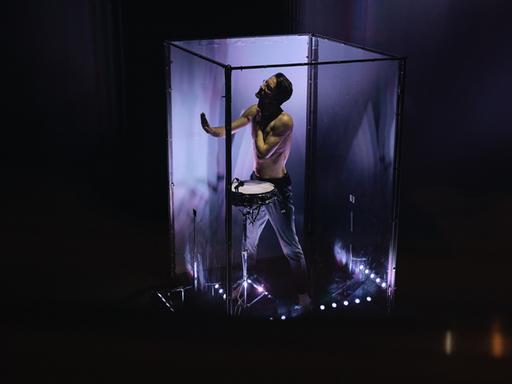 Ein Mann steht in einem lila ausgeleuchteten Glaskubus, wobei er eine Trommel an seinem Gürtel trägt und vorsichtig eine der vier Wände berührt. 