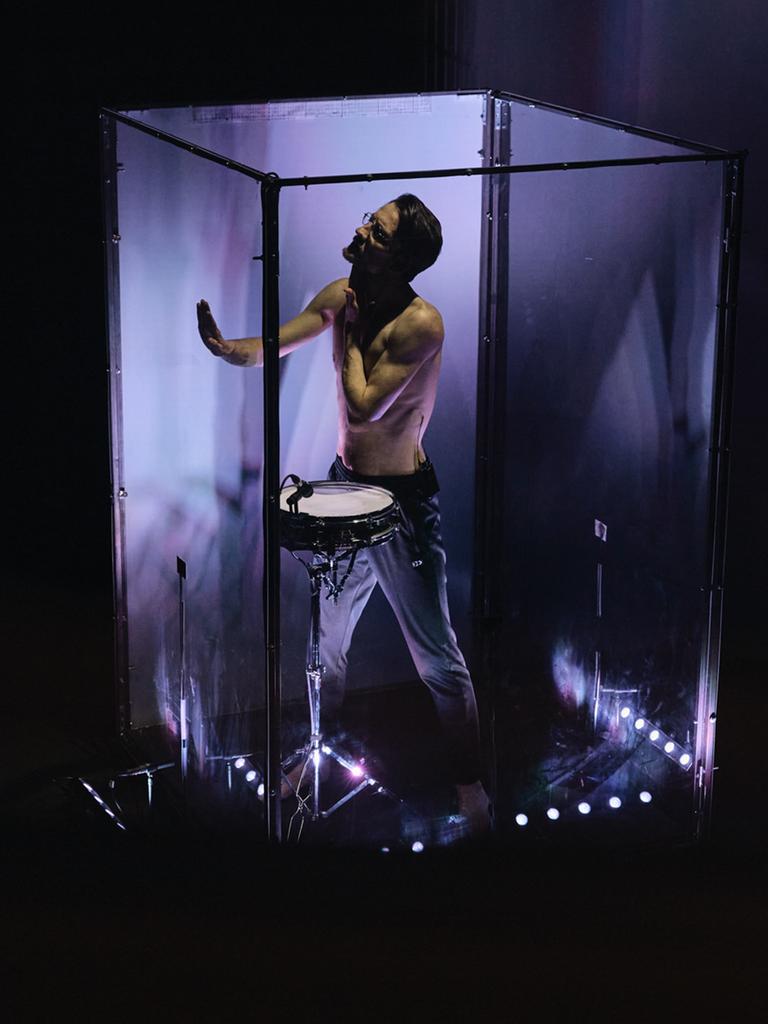 Ein Mann steht in einem lila ausgeleuchteten Glaskubus, wobei er eine Trommel an seinem Gürtel trägt und vorsichtig eine der vier Wände berührt. 