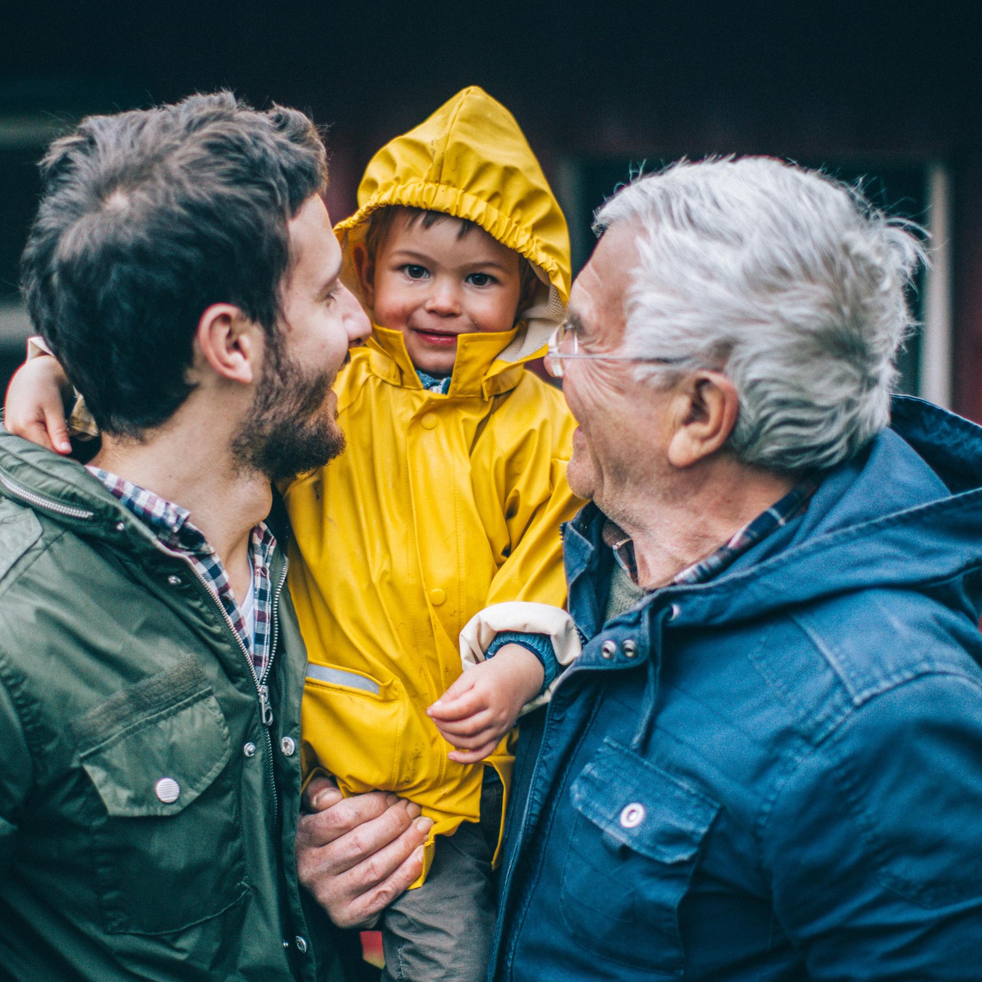 Ein Vater und ein Großvater tragen einen kleinen Jungen auf dem Arm - der Junge lacht in die Kamera, die beiden Erwachsenen lachen den Jungen an.

