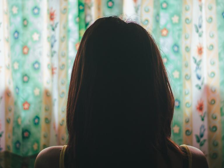 Eine Frau mit langen, braunen Haaren sitzt mit dem Rücken zur Kamera vor einem bunten Vorhang.
