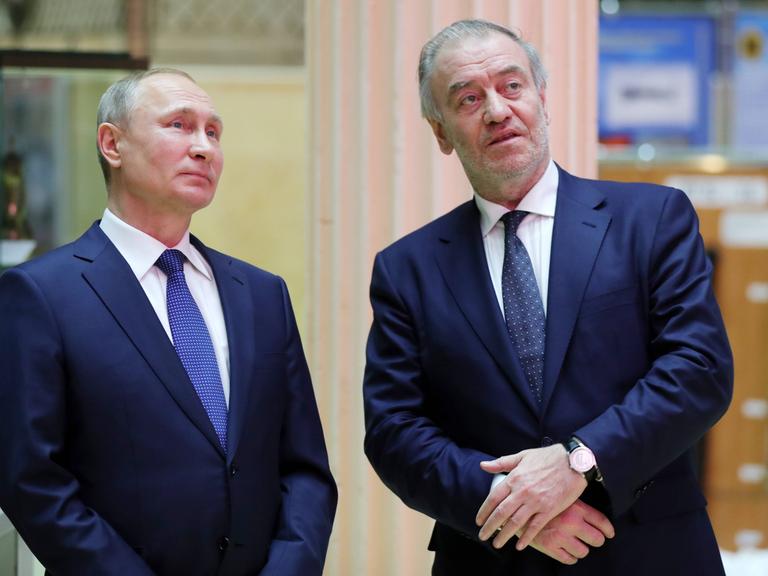 Der russische Präsident Wladimir Putin und der Dirigent Waleri Gergijew stehen nebeneinander. Putin auf der linken Seite, Gergijew auf der rechten. Sie tragen beide dunkelblaue Anzüge.