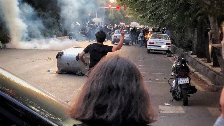 Ein junger Mann erhebt die Hand gegen Sicherheitskräfte im Hintergrund. Seitlich breitet sich Qualm auf der Straße aus Im Vordergrund steht eine junge Frau. Ein junger Mann erhebt die Hand gegen Sicherheitskräfte im Hintergrund. Seitlich breitet sich Qualm auf der Straße aus Im Vordergrund steht eine junge Frau.