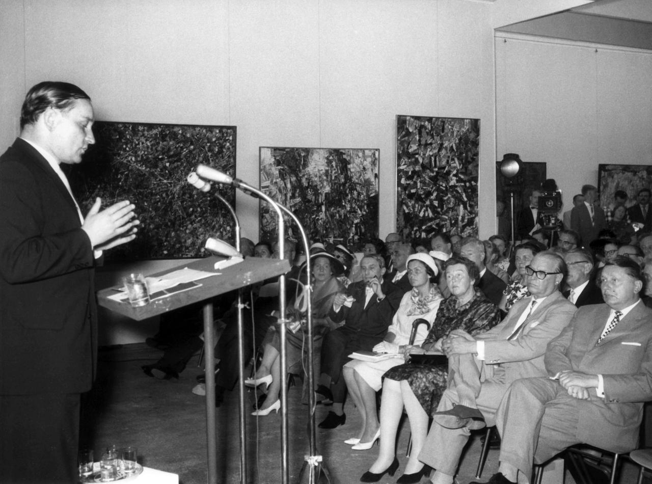 Ein Mann steht vor einem Podium und spricht zu einem sitzenden Publikum.