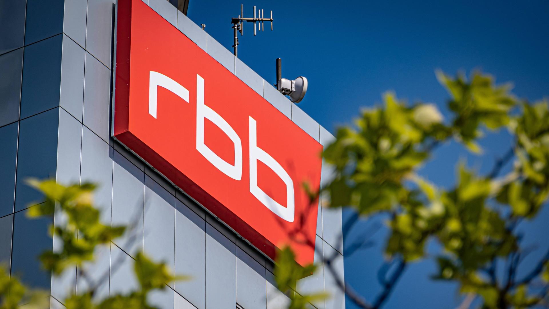 Das Logo und Gebäude des rbb im Hintergrund. Im Vordergrund unscharf Bäume. Das Logo und Gebäude des rbb im Hintergrund. Im Vordergrund unscharf Bäume.