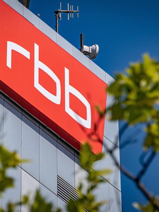 Das Logo und Gebäude des rbb im Hintergrund. Im Vordergrund unscharf Bäume.