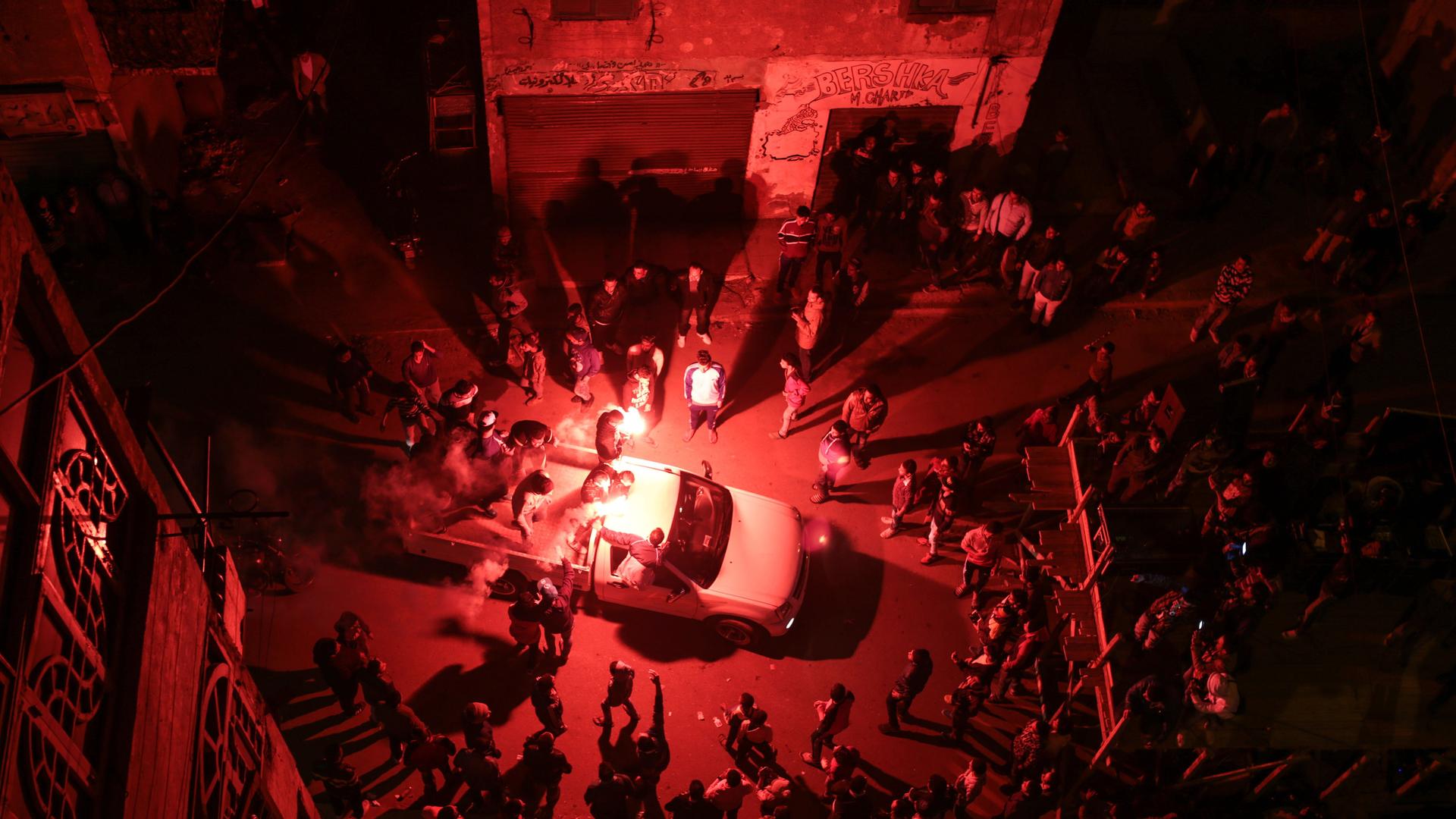 Luftaufnahme auf eine Ansammlung von Menschen bei Nacht von oben. In der Mitte ein Auto, darum sternenförmig tanzende Personen.