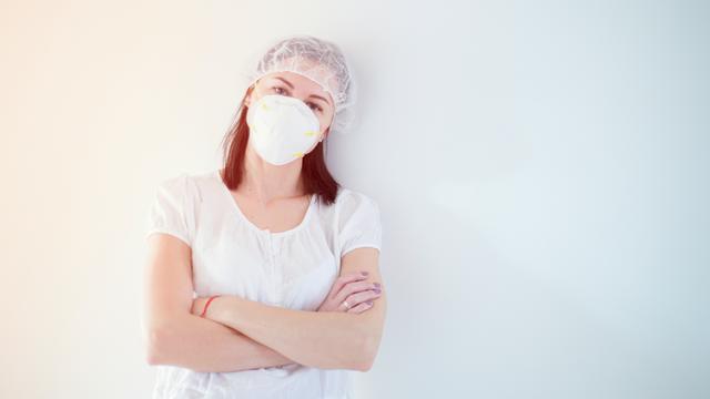 Portrait einer Frau in weißer Kleidung und mit Maske steht an eine weiße Wand gelehnt. 