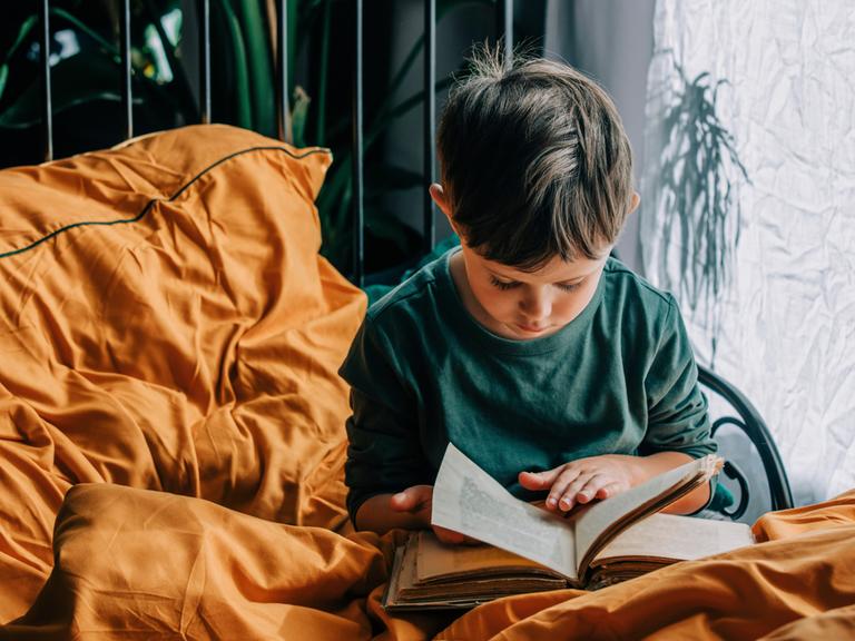 Ein Junge sitzt mit einem Buch auf dem Bett und liest.