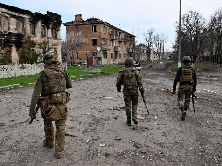 Drei bewaffnete Männer in Soldatenuniformen patrouillieren durch eine vom Krieg schwer beschädigte Kleinstadt.