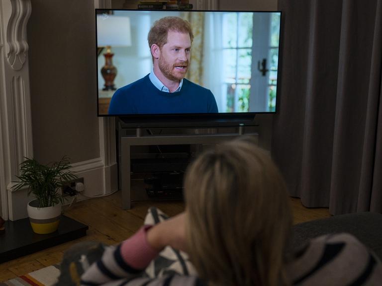 Eine Frau sitzt im Wohnzimmer vor dem Fernseher und schaut das Interview mit dem britischen Prinzen Harry an. 