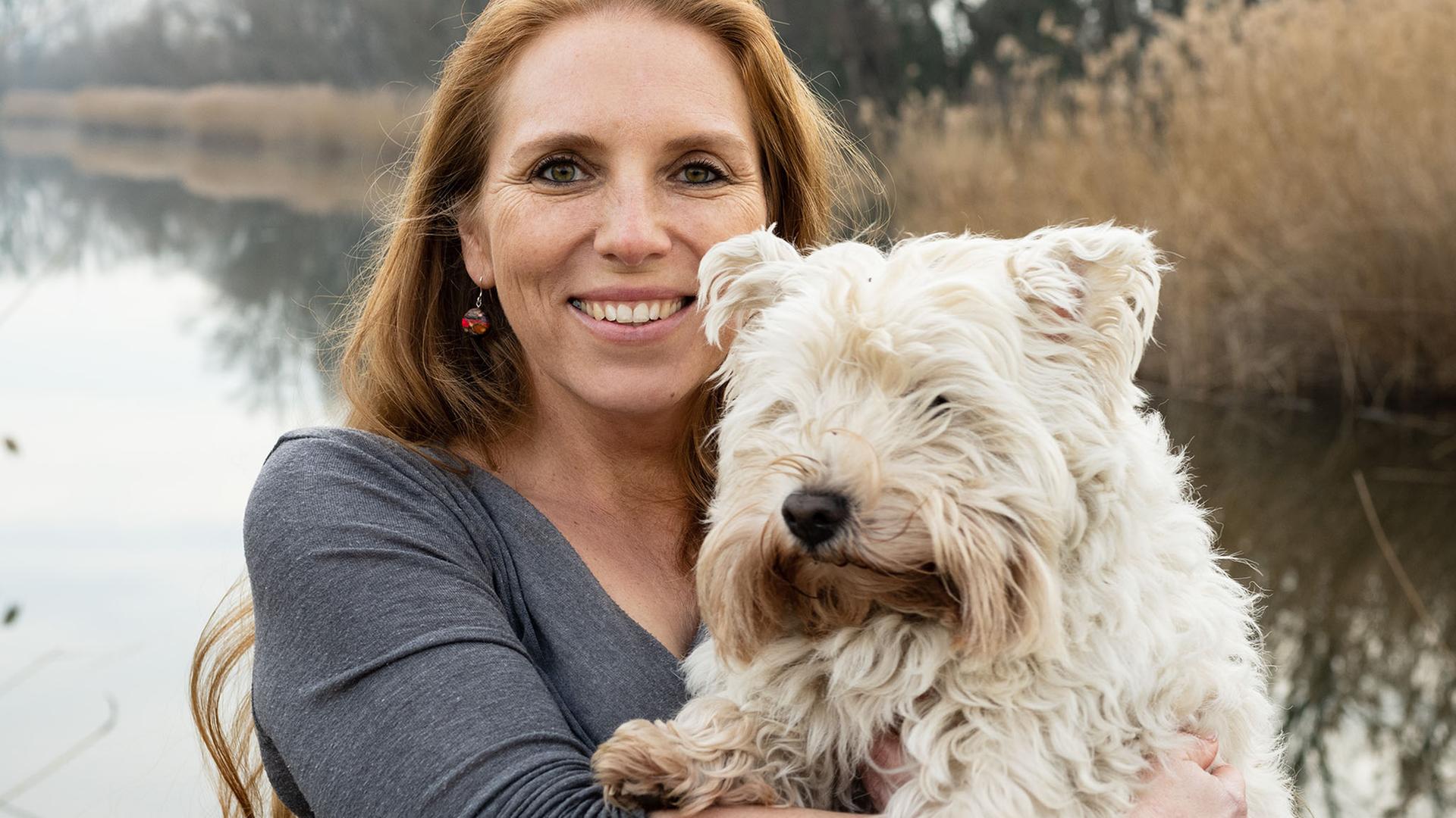 Die Biologin Angela Stöger trägt einen weißen, zotteligen Hund auf dem Arm, im Hintergrund ist ein Gewässer. Die Biologin Angela Stöger trägt einen weißen, zotteligen Hund auf dem Arm, im Hintergrund ist ein Gewässer.