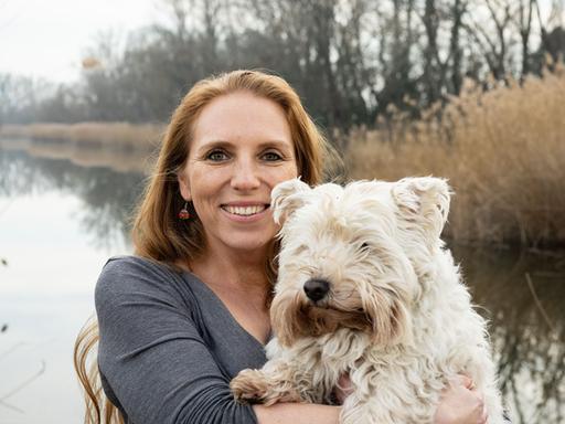 Die Biologin Angela Stöger trägt einen weißen, zotteligen Hund auf dem Arm, im Hintergrund ist ein Gewässer.