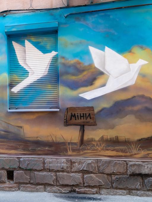 Ein Wandgemälde an einer Hausfassade zeigt mehrere weiße Tauben, die gen Himmel fliegen. Im Boden steckt ein Schild auf dem "Minen" steht. 