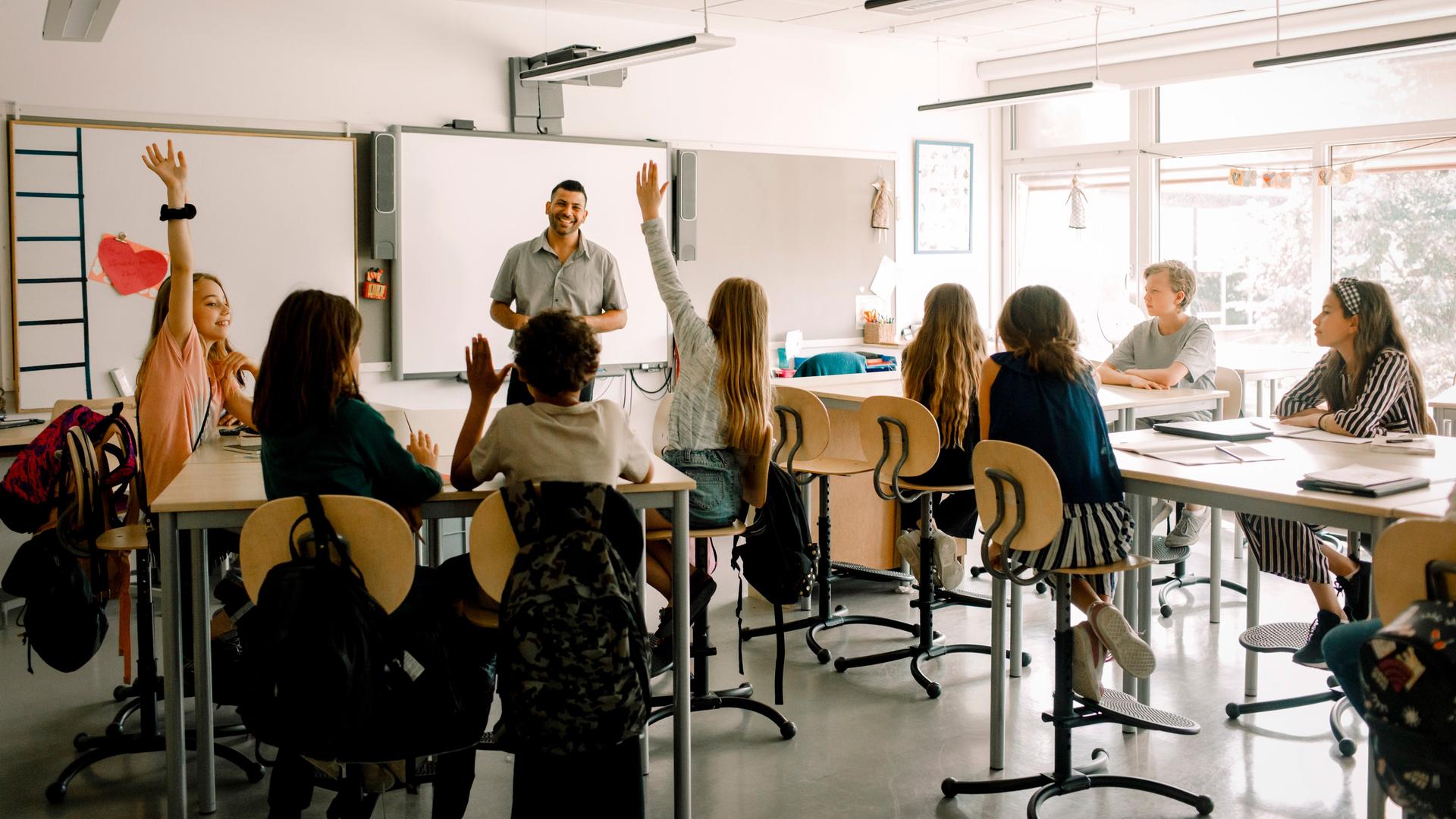 Schüler sitzen in einem Klassenraum. Vorn steht ein Mann mit dunklen, kurzen Haaren, der die Gruppe anlacht. Schüler sitzen in einem Klassenraum. Vorn steht ein Mann mit dunklen, kurzen Haaren, der die Gruppe anlacht.