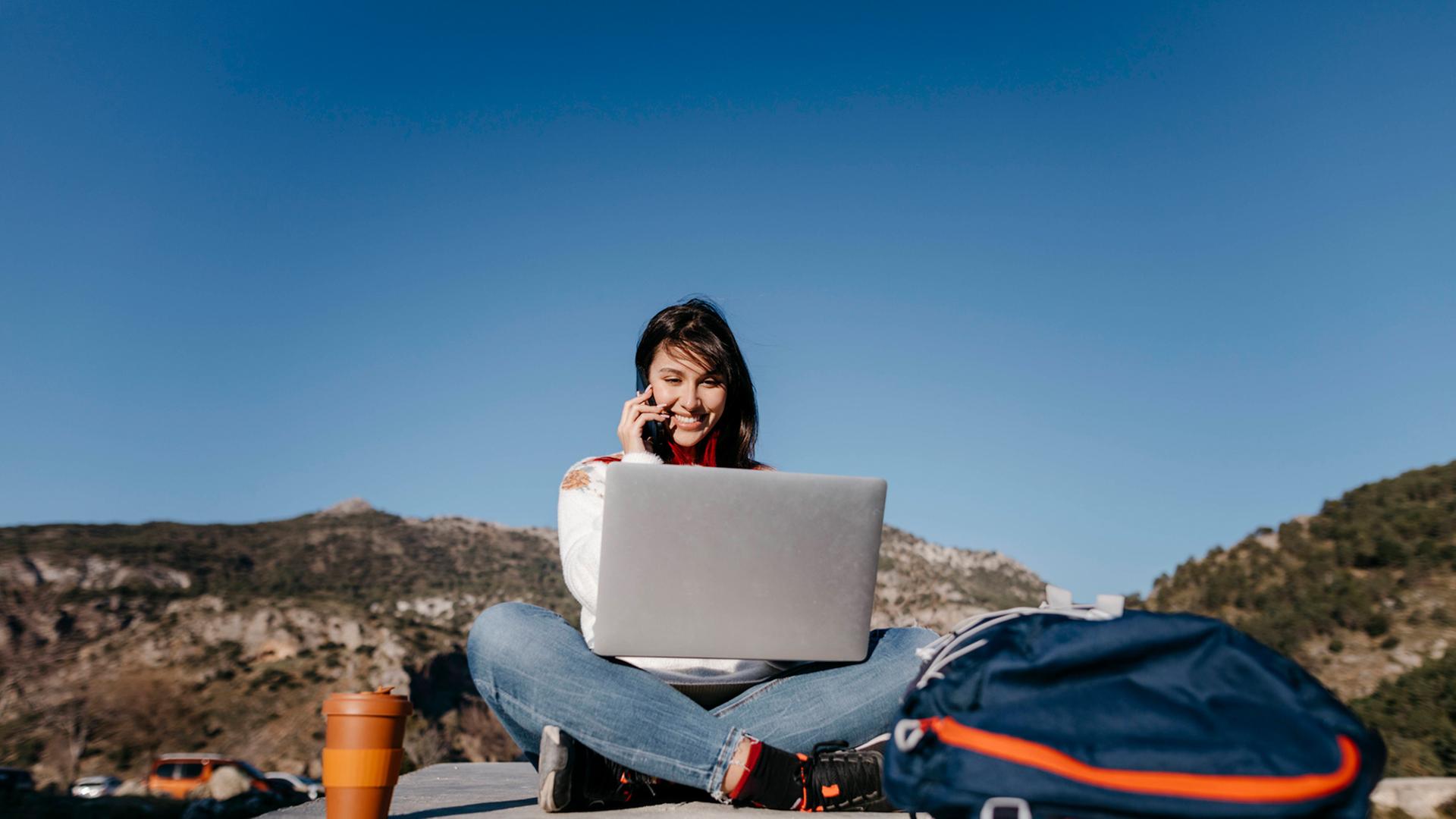 Arbeiten, wo andere Urlaub machen - für viele ein Traum. Zu sehen: Eine junge Frau sitzt auf einem Plateau (im Hintergrund sind Berge), hat einen Laptop auf dem Schoß und telefoniert mit einem Handy. Arbeiten, wo andere Urlaub machen - für viele ein Traum. Zu sehen: Eine junge Frau sitzt auf einem Plateau (im Hintergrund sind Berge), hat einen Laptop auf dem Schoß und telefoniert mit einem Handy.