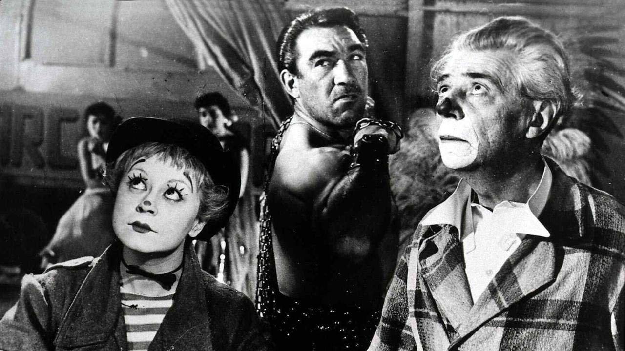Drei Menschen blicken nach oben aus dem Bild heraus. Vermutlich befinden sie sich in einem Zirkus und schauen Richtung Kuppel. Die Frau links und der Mann rechts sind geschminkt wie Clowns. Der Mann in der Mitte steht etwas Abseits, er schaut finster. Drei Menschen blicken nach oben aus dem Bild heraus. Vermutlich befinden sie sich in einem Zirkus und schauen Richtung Kuppel. Die Frau links und der Mann rechts sind geschminkt wie Clowns. Der Mann in der Mitte steht etwas Abseits, er schaut finster.