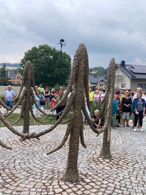 Sechs Fichten aus Bronze stehen in einem Kreis und halten sich freundschaftlich an den Ästen. Erst auf den zweiten Blick sieht man ihre Gesichter.