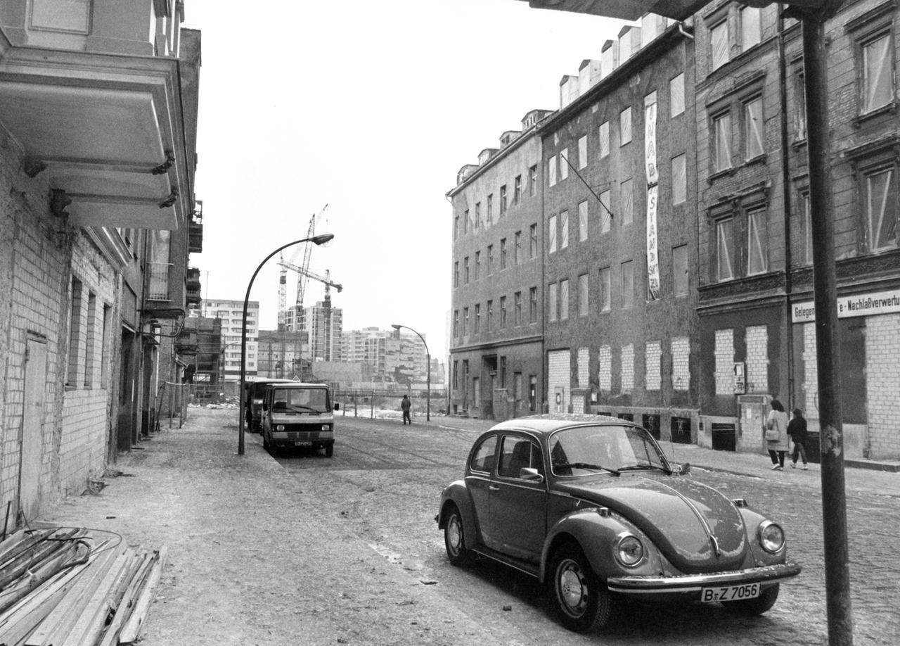 Schwarz-weiß-Aufnahme von einer Straße in Berlin-Kreuzberg Anfang der 1980er-Jahre. Auf der linken Seite werden Altbauten restauriert, auf der rechten stehen Abrisshäuser. Im Vordergrund parkt ein VW Käfer.