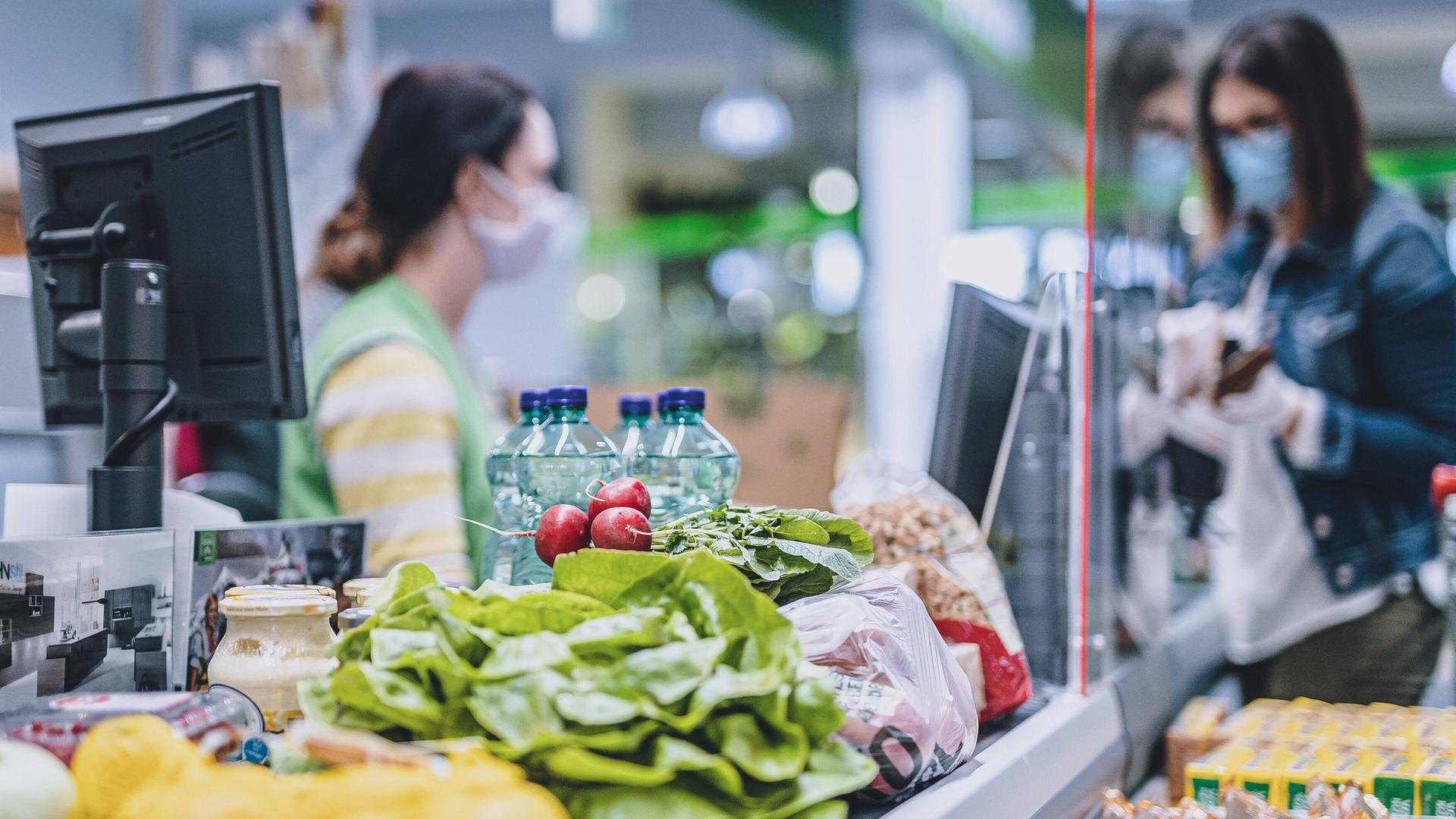 Eine Frau trägt eine Schutzmaske und Handschuhe bei ihrem Einkauf in einem Supermarkt, während sie an der Kasse bezahlt.