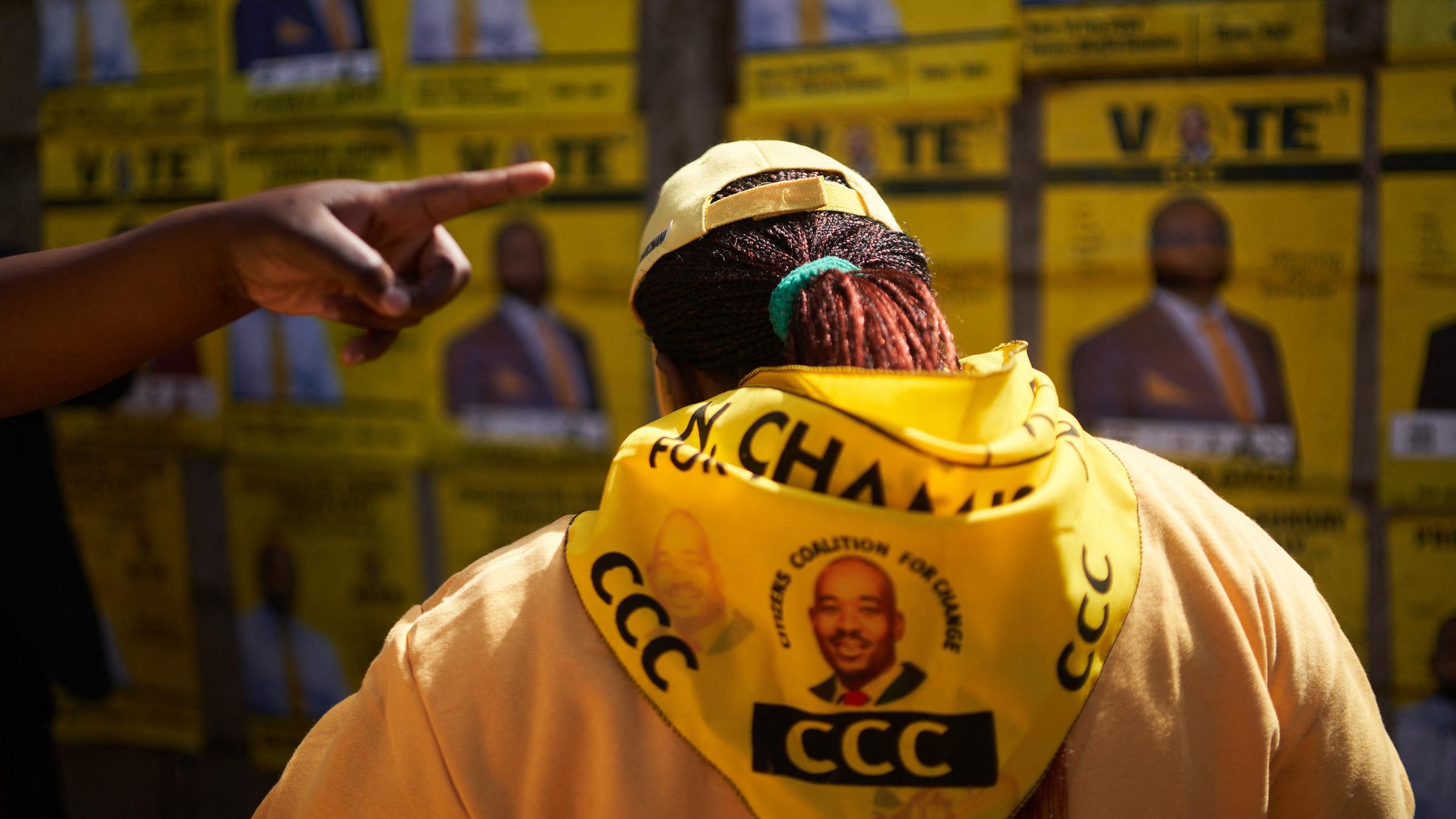 Ein Unterstützer der Oppositionspartei trägt eine gelbe Jacke mit einem aufgedruckten Porträt des Kandidaten. Im Hintergrund sind verschwommen Wahlplakate zu sehen.