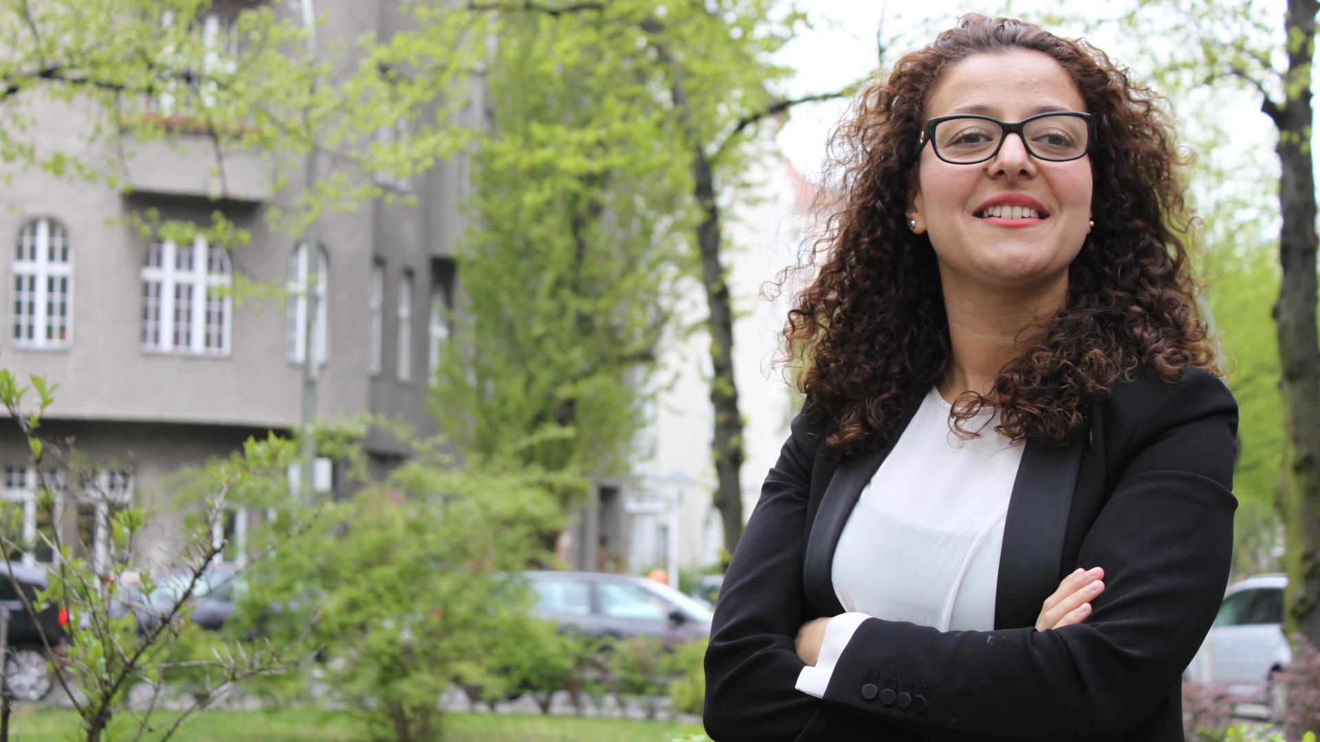 Die Sozialmanagerin Jouanna Hassoun ist Geschäftsführerin des Vereins Transaidency. Sie steht auf einer Grünfläche vor einem Haus in Berlin und verschränkt die Arme.
