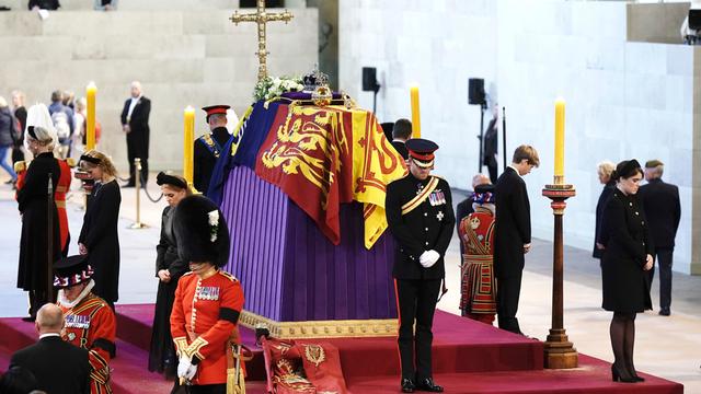Uniformierte Männer und Frauen in schwarzer Trauerkleidung umstehen auf eine, rotem Podest einen Sarg, der mit einer Flagge, Blumen und einem goldenen Kreuz geschmückt ist.