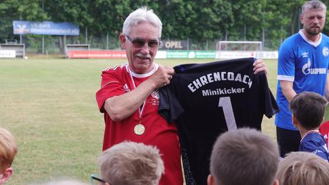 Ein Mann mit weißen, kurzen Haaren im Fußballtrikot und Medaille um den Hals hält vor ein paar Kindern ein T-Shirt mit dem Aufdruck "Ehrencoach Minikicker 1" hoch.