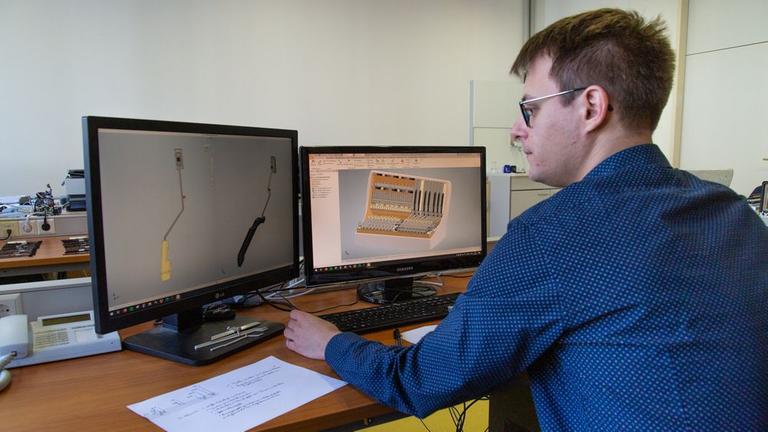 Ein junger Mann mit Brille und kurzen braunen Haaren sitzt vor zwei Computerbildschirmen. Darauf sieht er links zwei Hebel, und rechts einen weißen Kasten, mit vielen Bauteilen drin. Ein junger Mann mit Brille und kurzen braunen Haaren sitzt vor zwei Computerbildschirmen. Darauf sieht er links zwei Hebel, und rechts einen weißen Kasten, mit vielen Bauteilen drin.