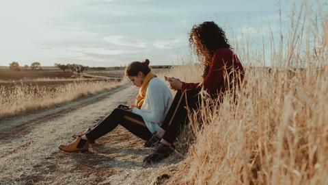 Zwei Frauen sitzen mit Mobiltelefonen in der Hand an einem Feldweg. 