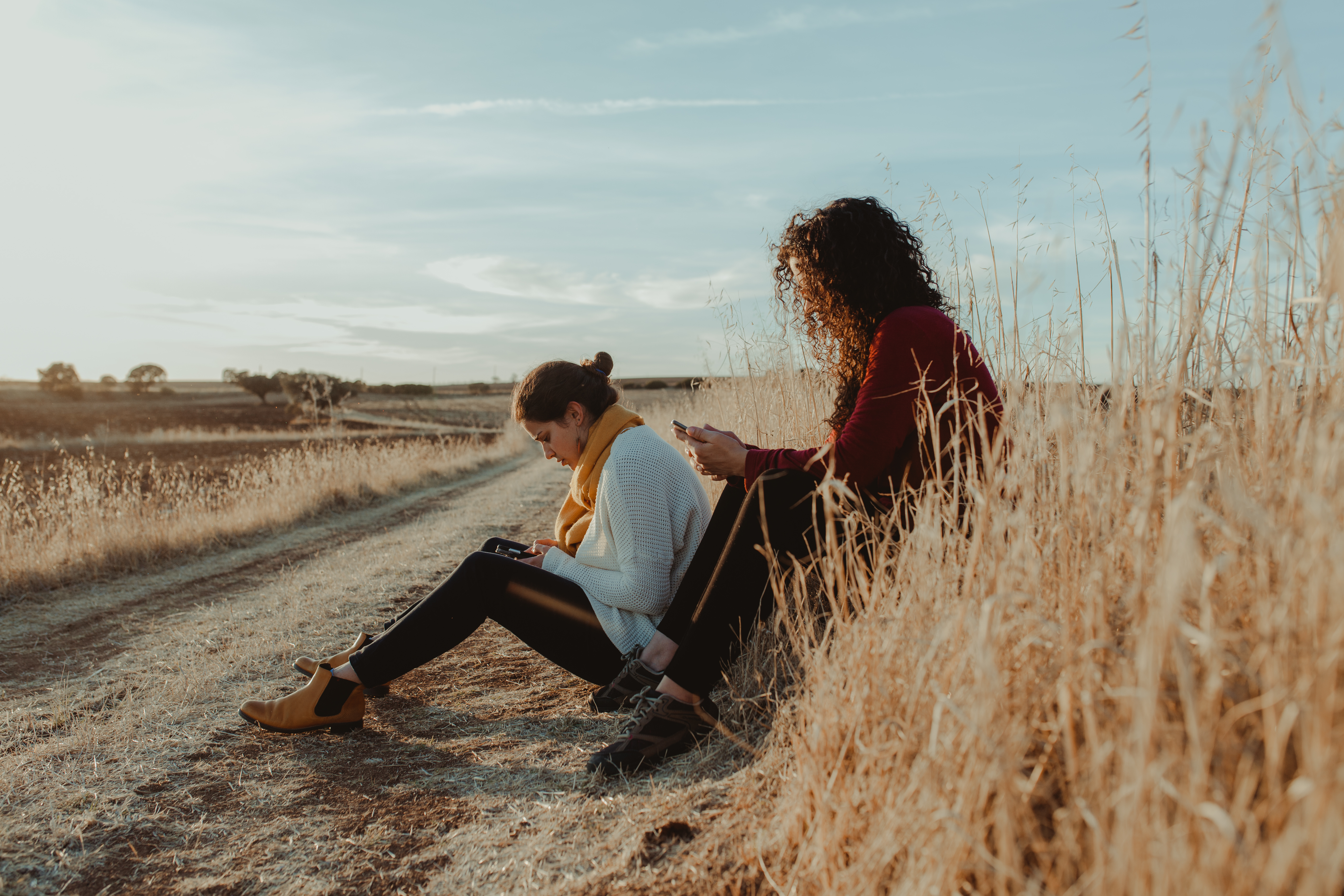 Zwei Frauen sitzen mit Mobiltelefonen in der Hand an einem Feldweg. 