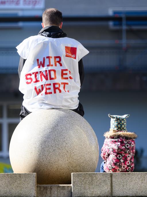 Ein Erzieher sitzt auf einem Stein. Auf seinem Rück steht "Wir sind es wert". Neben ihm sitzt ein kleines Kind.