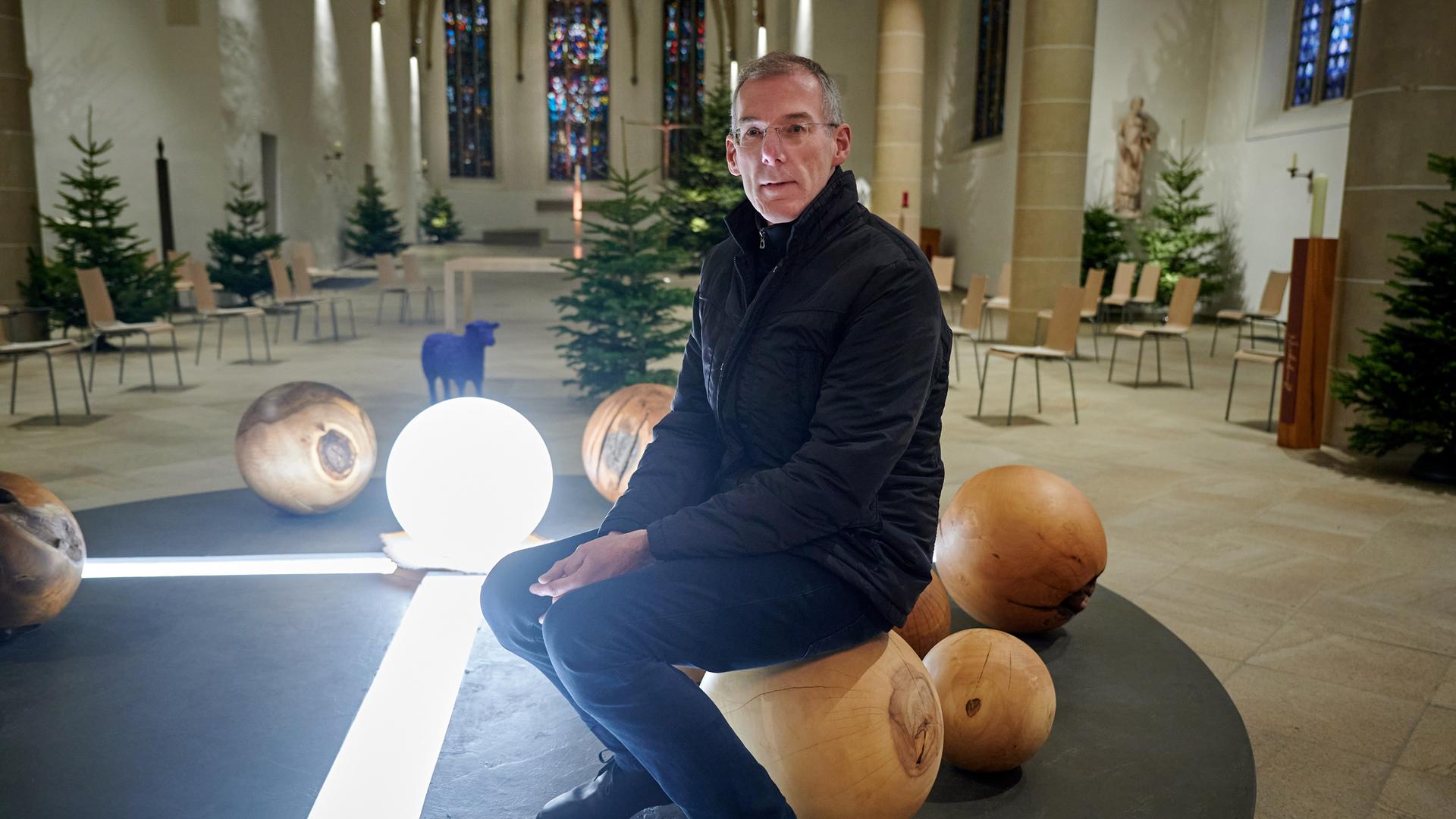 Bernd Mönkebüscher, in schwarzem Anorak und schwarzer Hose, sitzt in einem hellen Kirchenraum auf einer Holzkugel, die zu einer Lichtinstallation gehört. Bernd Mönkebüscher, in schwarzem Anorak und schwarzer Hose, sitzt in einem hellen Kirchenraum auf einer Holzkugel, die zu einer Lichtinstallation gehört.