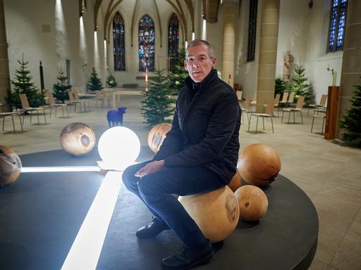 Bernd Mönkebüscher, in schwarzem Anorak und schwarzer Hose, sitzt in einem hellen Kirchenraum auf einer Holzkugel, die zu einer Lichtinstallation gehört.