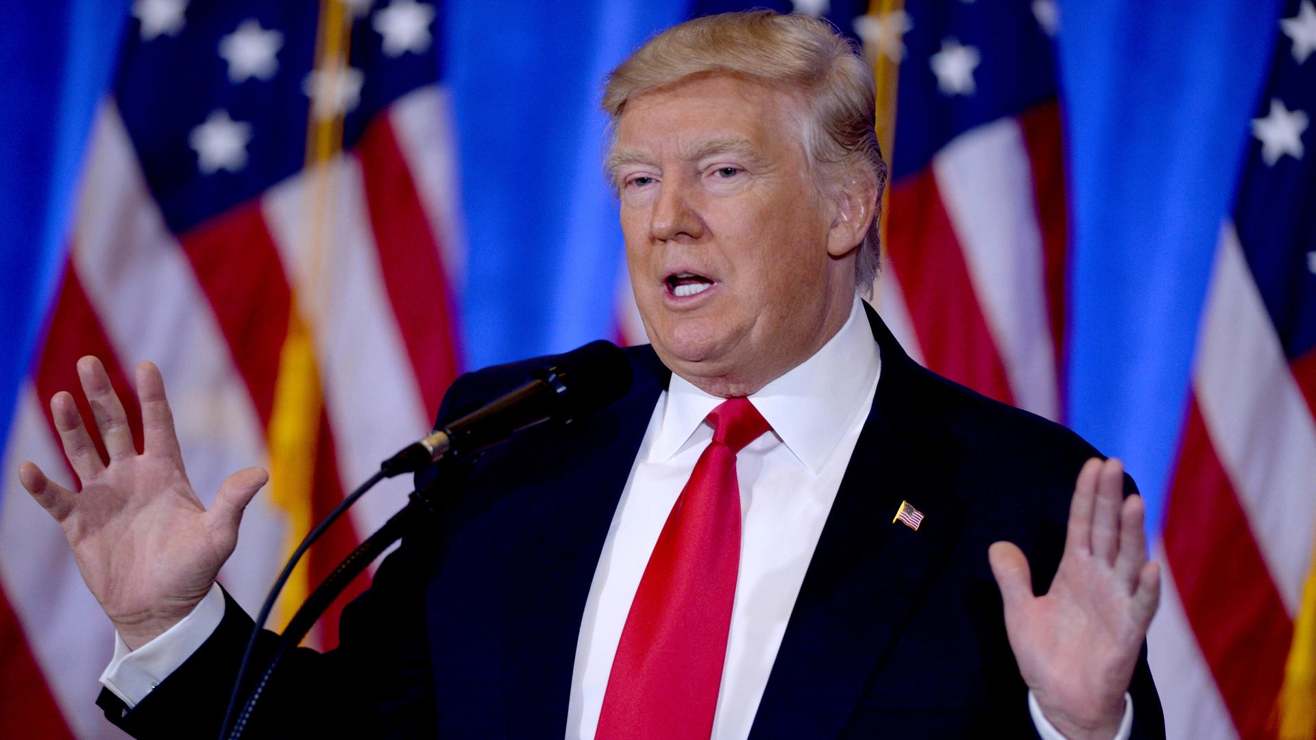 Das Foto zeigt Donald Trump. Er steht hinter einem Rednerpult und streckt die Hände nach oben.