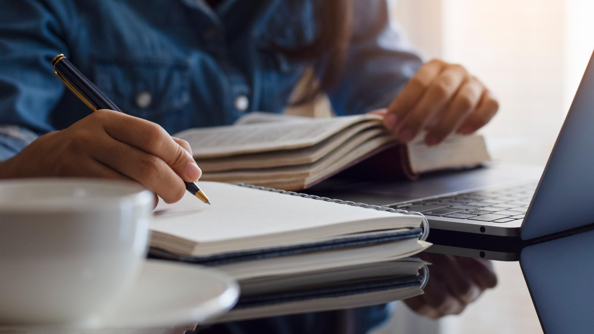 Eine Frau liest ein Buch und macht sich daneben Notizen in einem Heft mit leeren Seiten. Eine Frau liest ein Buch und macht sich daneben Notizen in einem Heft mit leeren Seiten.