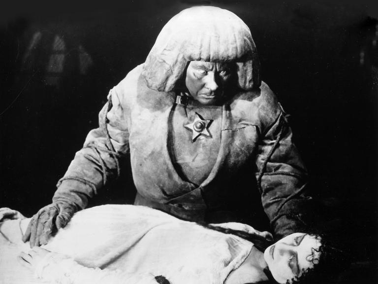 Filmszene aus "Der Golem, wie er in die Welt kam" von 1920. Darin zu sehen ist Paul Wegener als Golem, wie er sich über eine liegende Frau beugt.